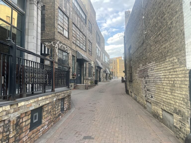 Brick-paved Fawkes Alley lined with old auto buildings, striped awnings and string lights