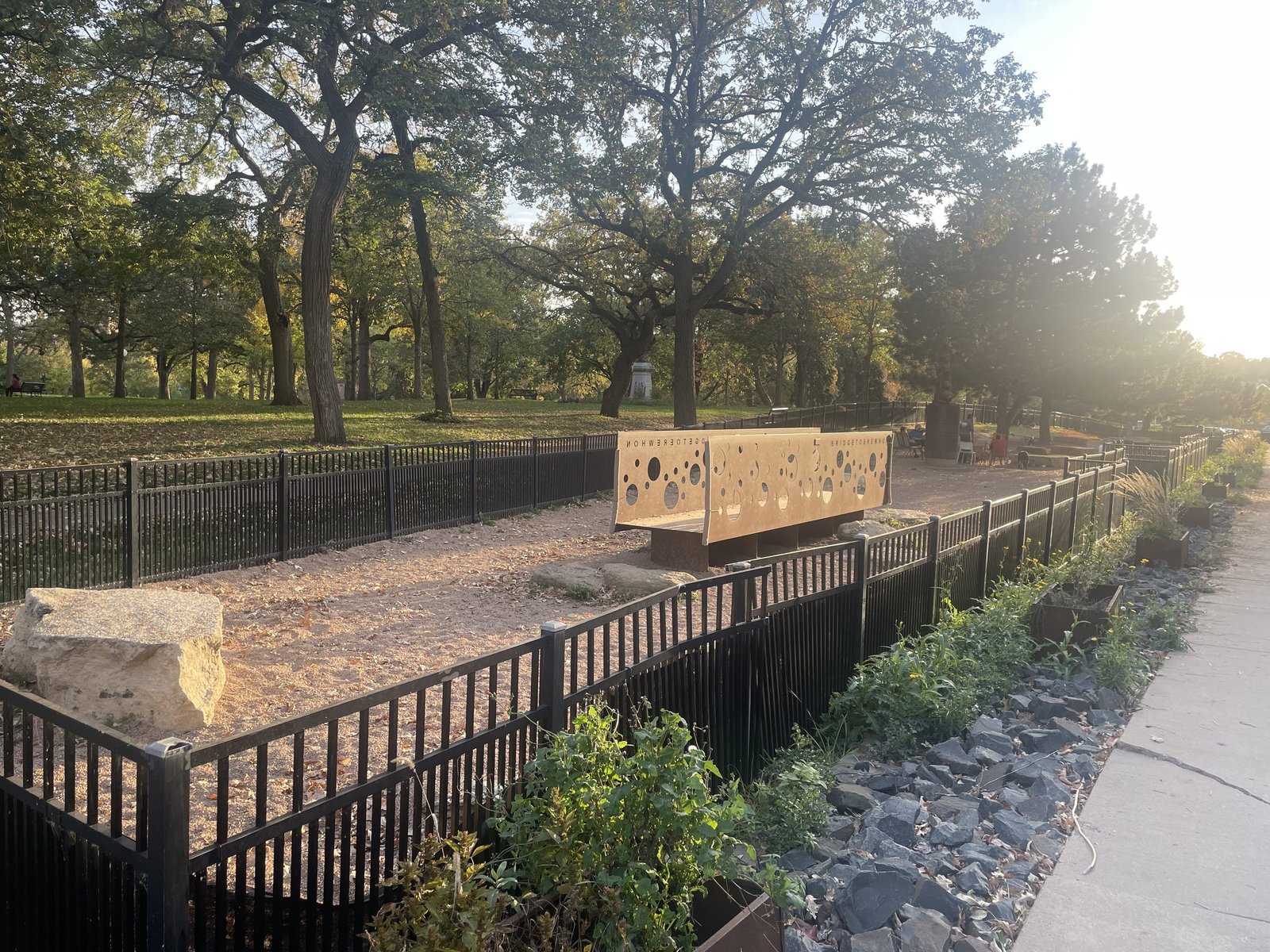 Fenced dog park in Loring Park with a perforated sculptural bridge and large oak trees